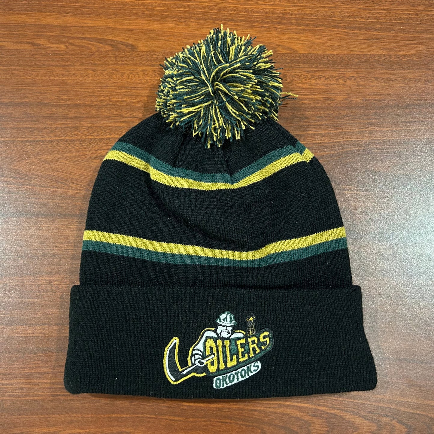 Black toque with green and gold stripes and pom sitting on a brown table. The toque features a fully embroidered Okotoks Oilers primary logo on the cuff.