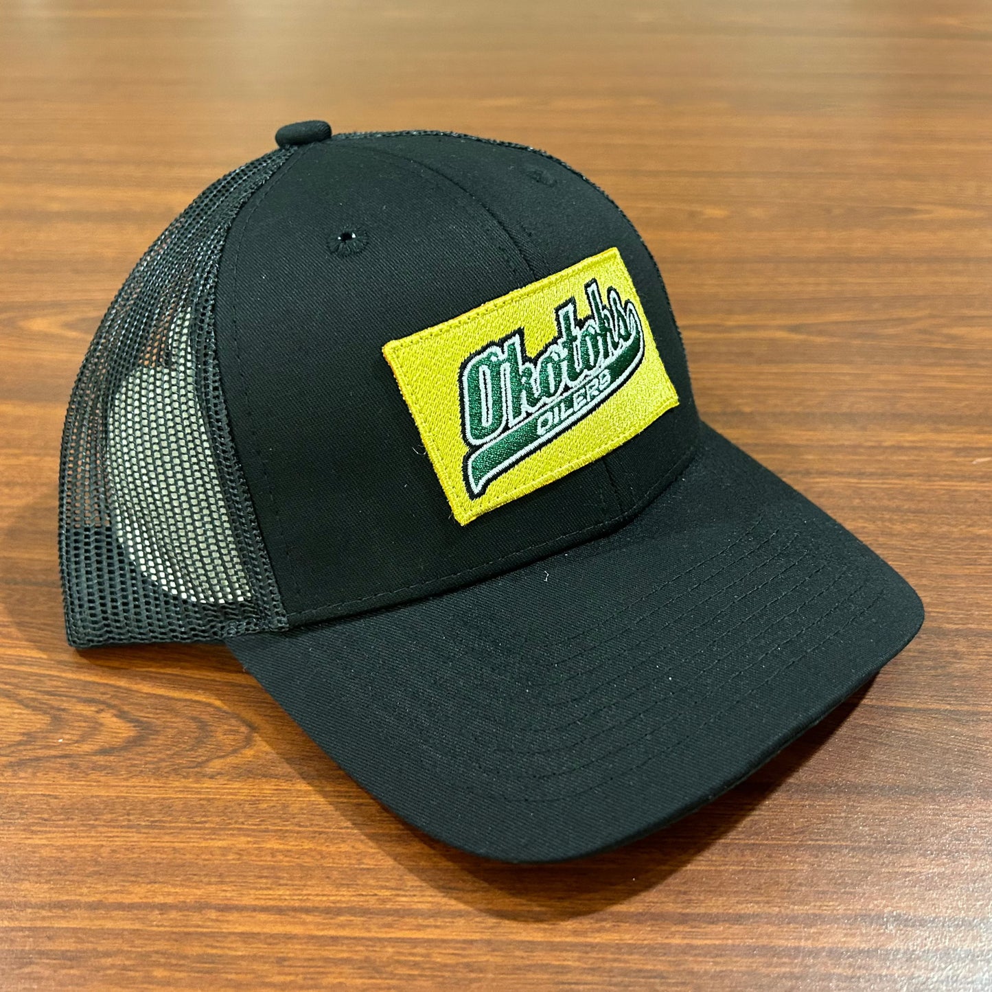Angled view of a black baseball cap with a gold patch featuring the Okotoks Oilers' green script logo.