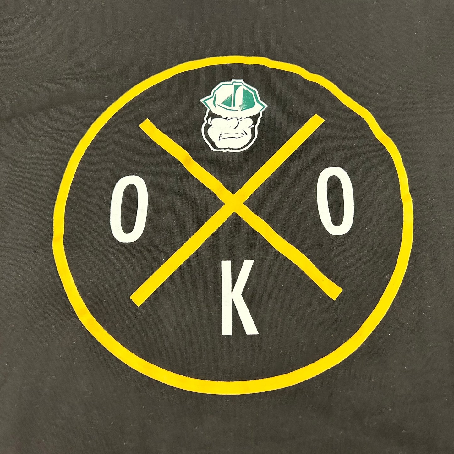 Cropped view of a black T-Shirt with a circular design on the chest featuring the letters OKO and the Oilers' Rigger head logo.