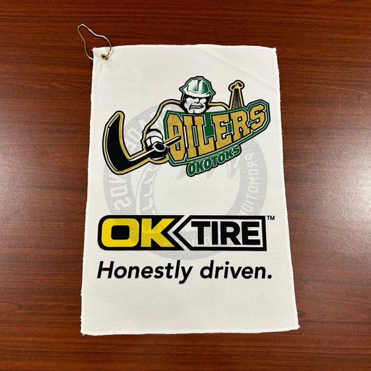 White golf towel with the Oilers' primary logo and the logo of OK Tire. The towel can be attached to a golf bag with the gold clip in the top-left corner.