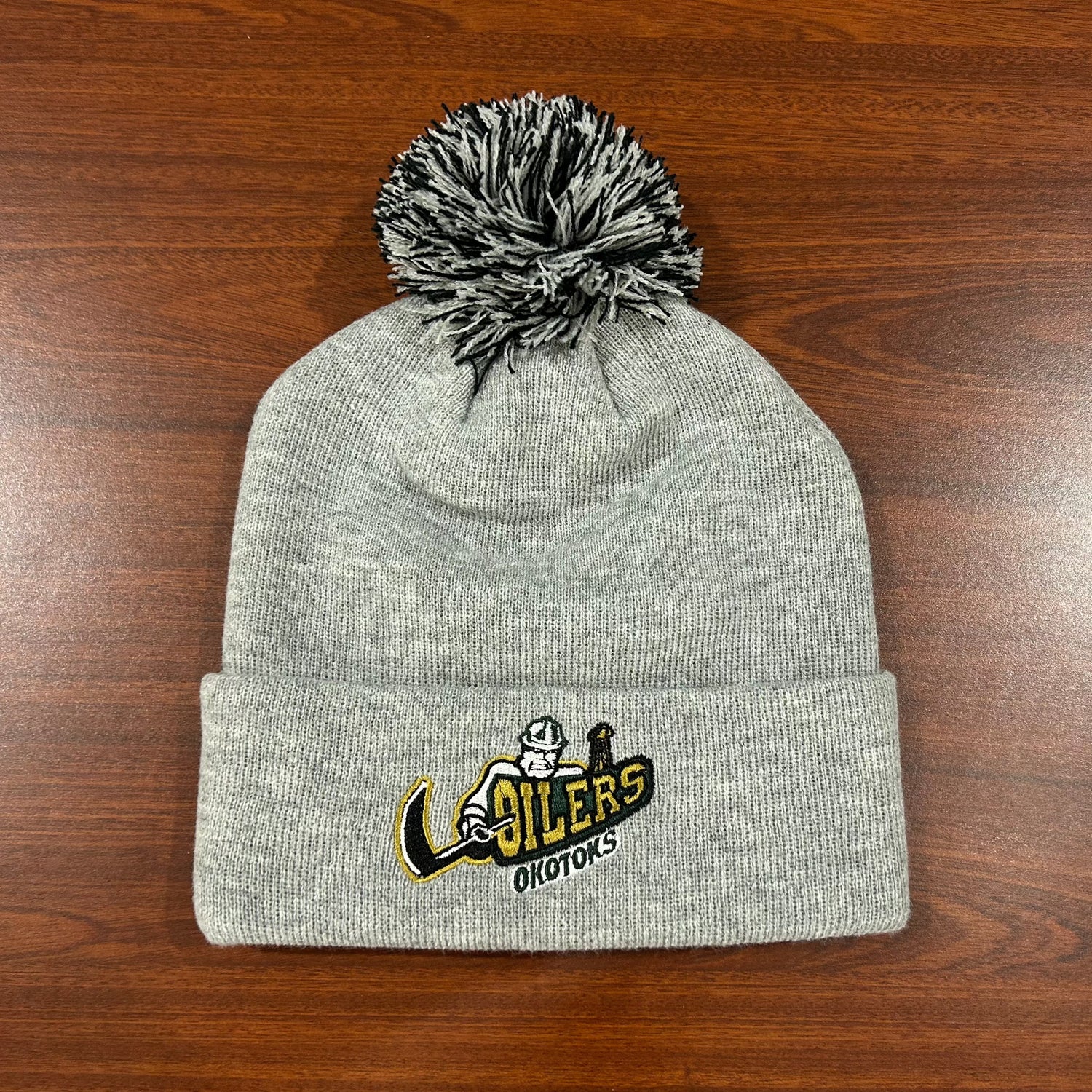 Grey toque with black and grey pom sitting on a brown table. The toque features a fully embroidered Okotoks Oilers primary logo on the cuff.