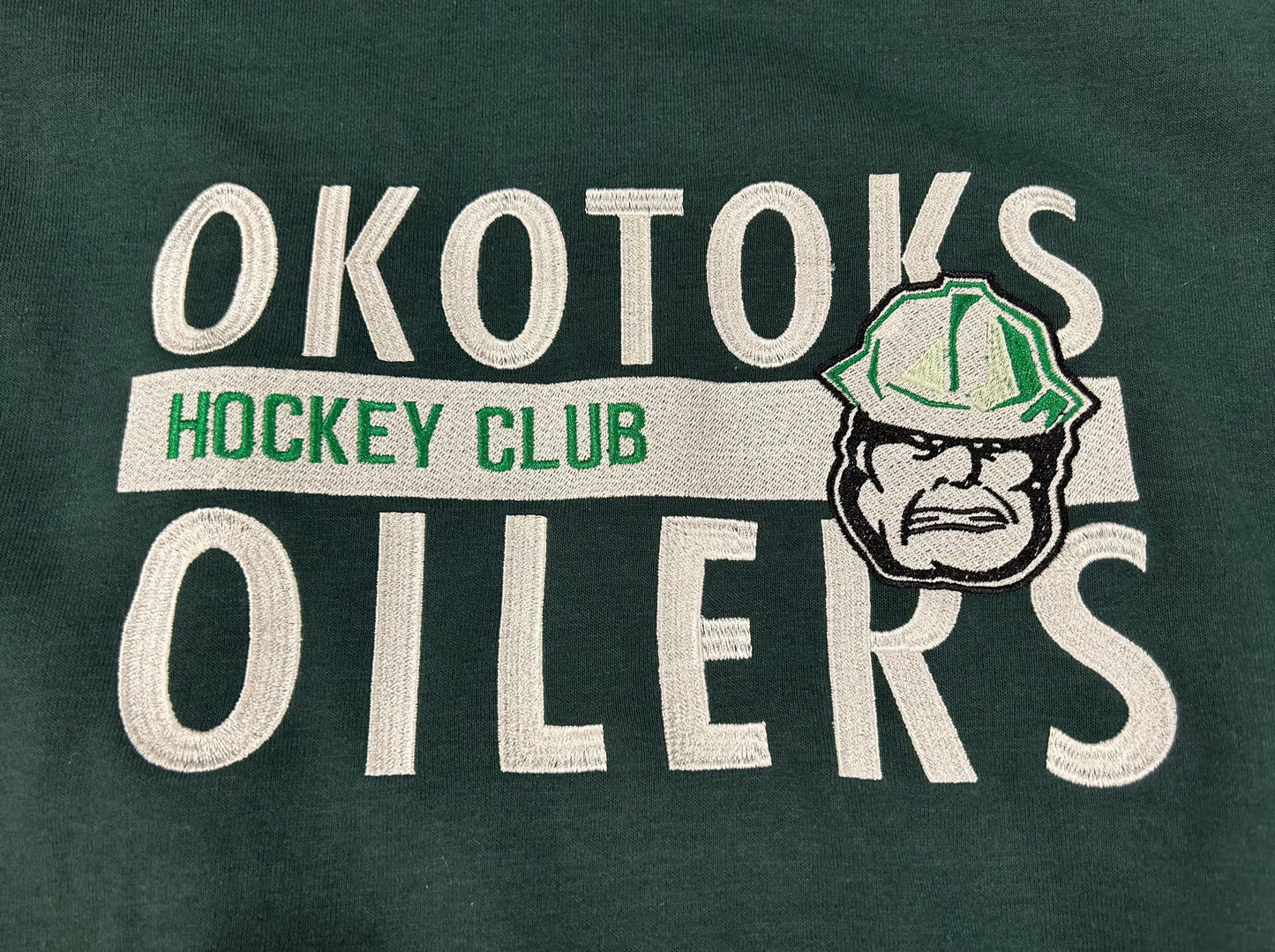 Cropped view of green hoodie with OKOTOKS OILERS HOCKEY CLUB and the Okotoks Oilers' Rigger head logo embroidered on the chest.