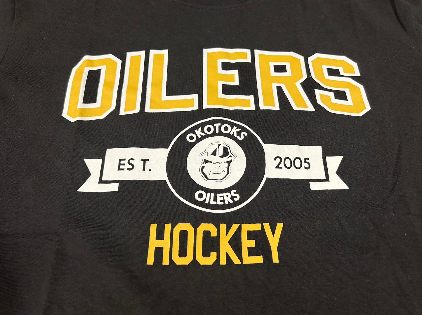 Cropped view of a black T-Shirt with OILERS HOCKEY printed on the chest. Between those two words is a small banner with the Oilers' Rigger head logo and text reading "OKOTOKS OILERS EST. 2005".